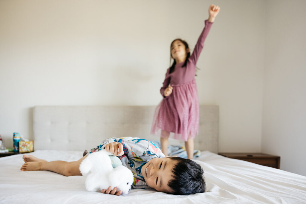 Older sister is jumping on the bed while younger toddler brother laughs.