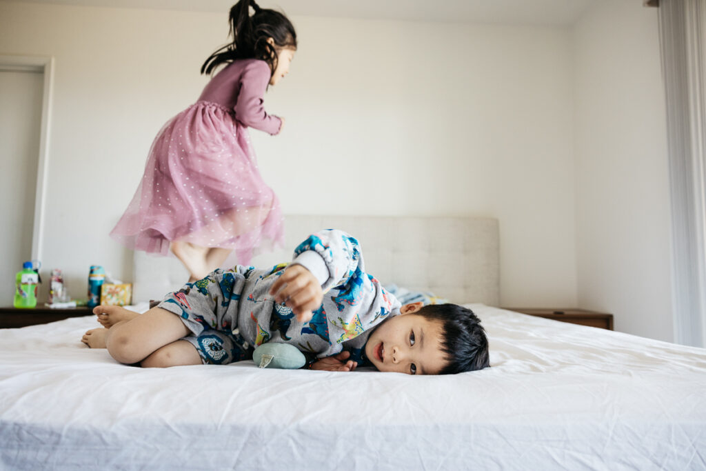 Older sister is jumping on the bed while younger brother watches.