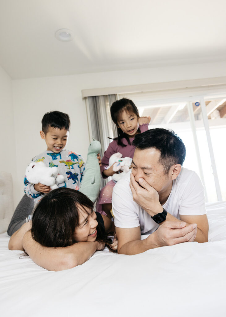 Mom and dad are laughing hysterically on the bed while their kids watch.