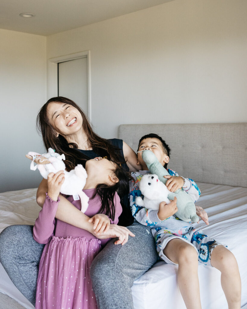 Mom is laughing while holding her young kids on the bed.