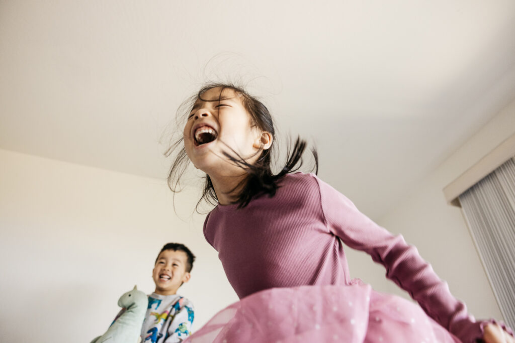 Sister is jumping on the bed and laughing while her brother laughs.
