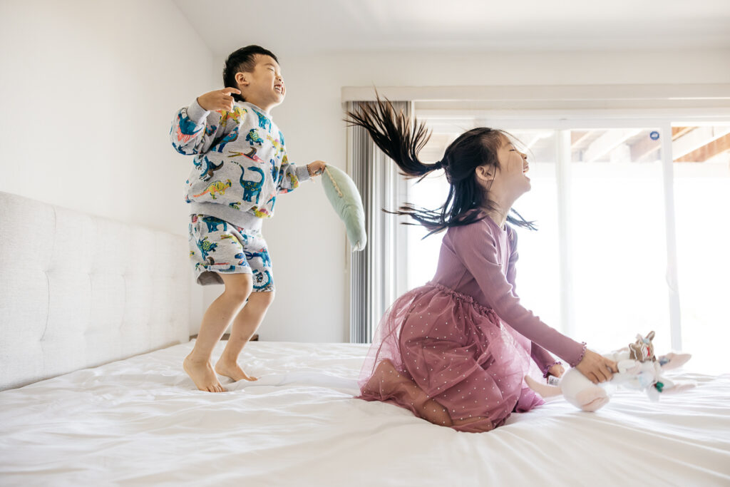 Young siblings are laughing while jumping on the bed during a family photo session at home in Oakland.