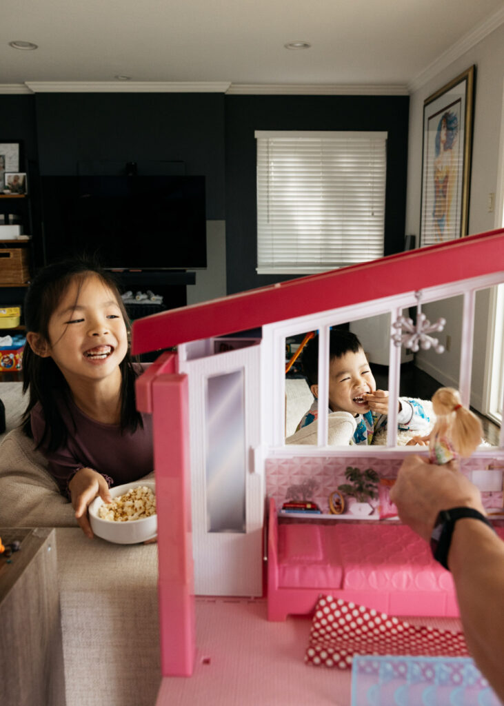 Sibling brother and sister are laughing at dad while he plays with Barbie during a family photo session.