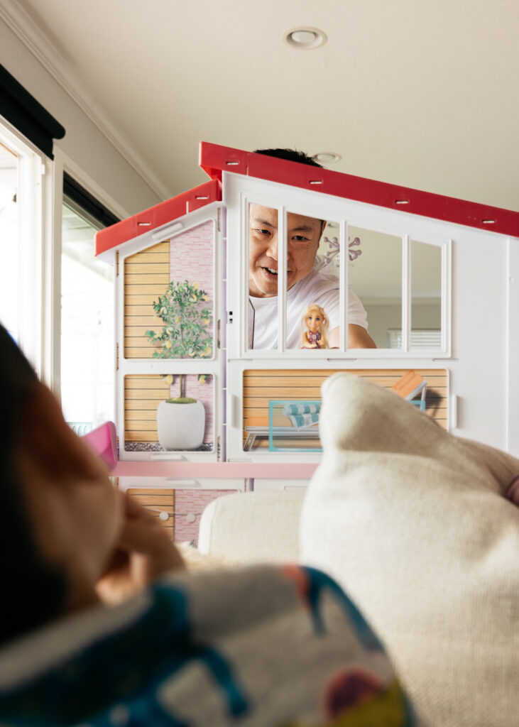 Dad is playing with Barbie in the dollhouse while his kids laugh.