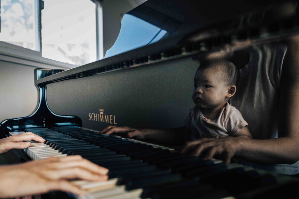 A reflection of a baby girl is shown at the piano while she sits in her mom's lap as she plays.