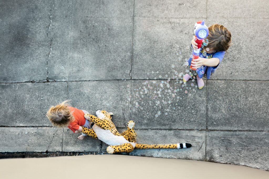 Young toddler boy drags a large stuffed cheetah across the driveway while his older brother blows bubbles.