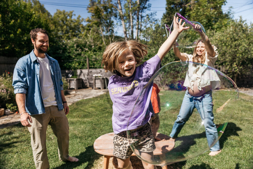 Boy is jumping to catch a large bubble while mom is creating them with a large wand.