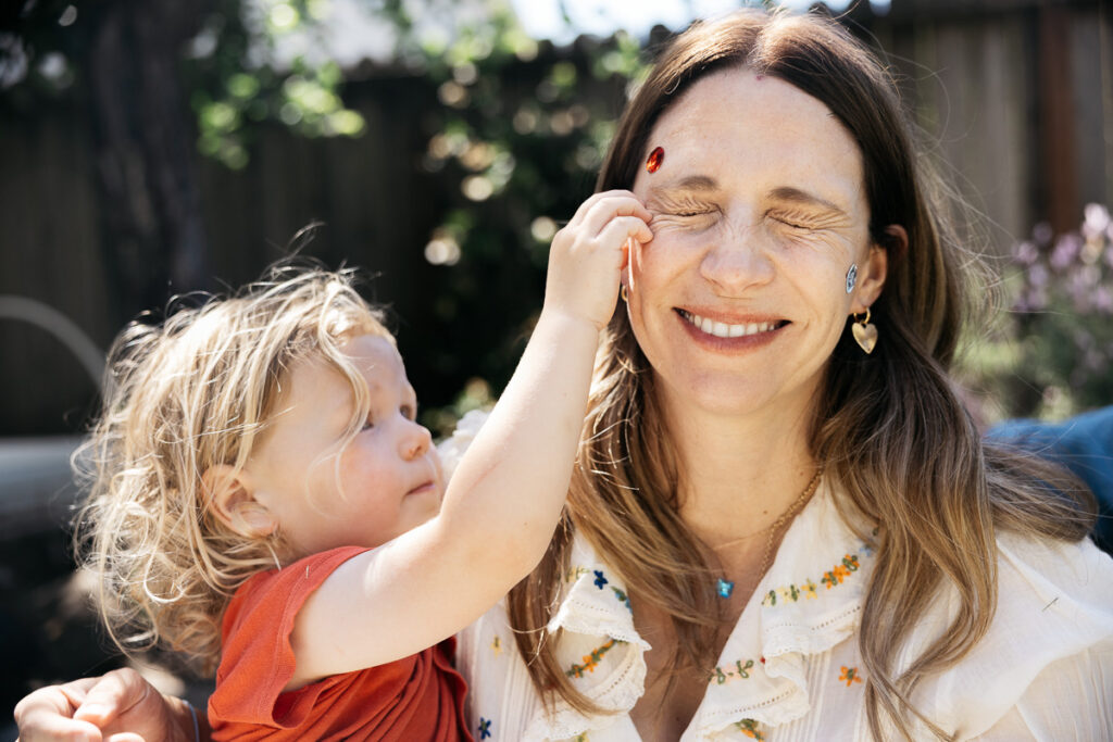 Toddler boy is putting stickers on his mom's face while she closes her eyes and laughs.