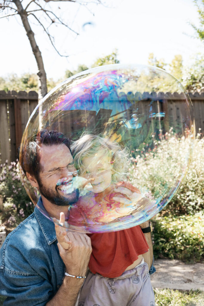 Dad and young toddler son is squinting their eyes as they anticipate a bubble to burst as it floats by their faces.